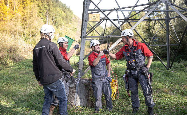 Vier junge Männer in APG-Schutzkleidung vor einem Strommast auf einer Wiese. Sie lachen und geben sich High-five.